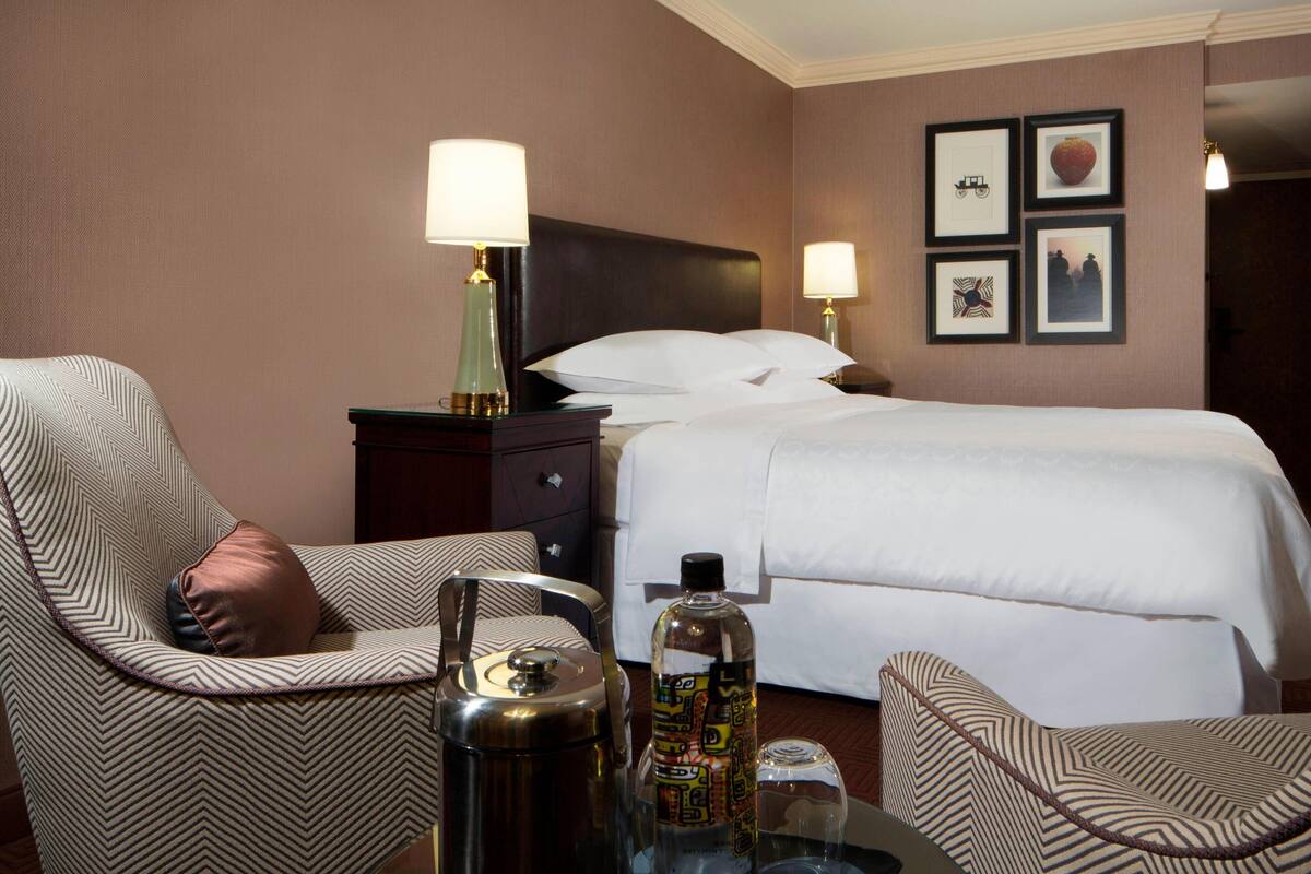 A hotel room with a bed, two chairs, a nightstand with a lamp, a bottle, and a thermos.