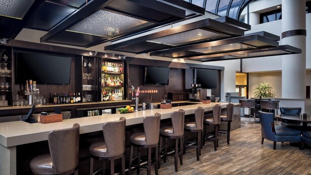 A modern bar area with a long counter, multiple bar stools, and a television screen. - Sheraton DFW Airport Hotel (Irving)