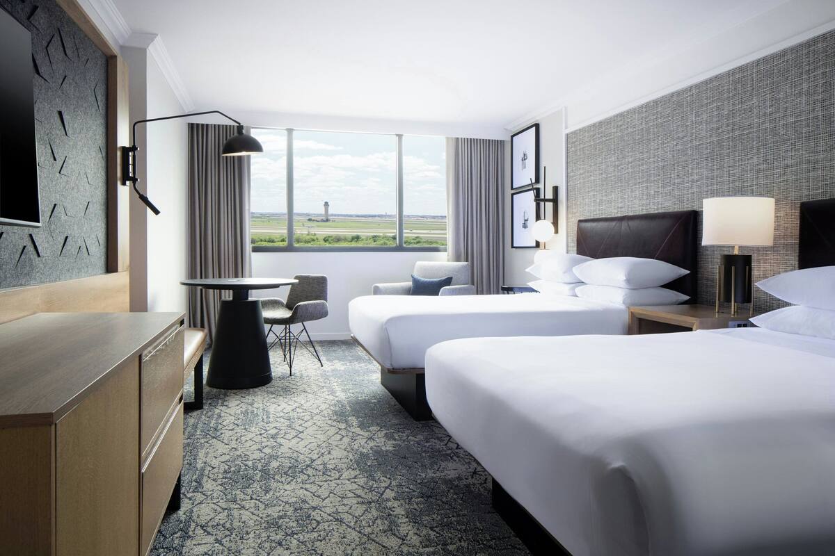 A hotel room with two beds, a desk, a chair, a TV, and a large window with a view of the countryside.