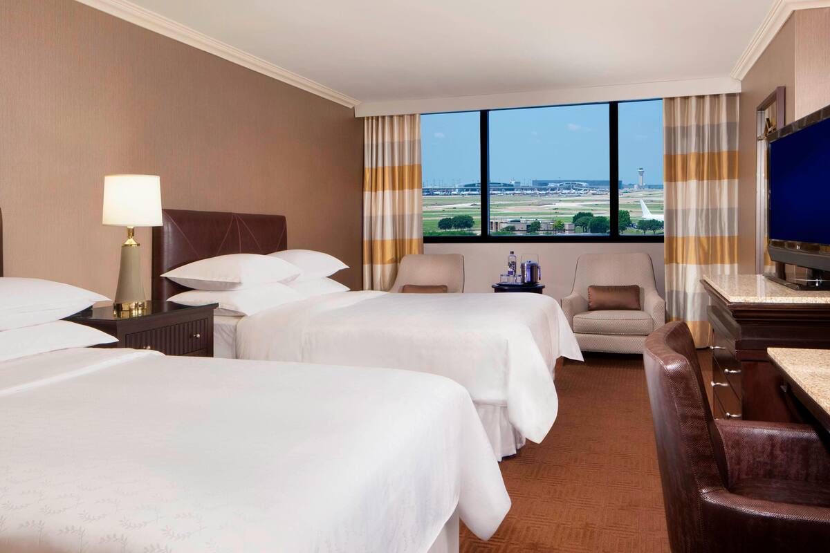 A hotel room with two beds, a TV, a desk, and a view of the airport.