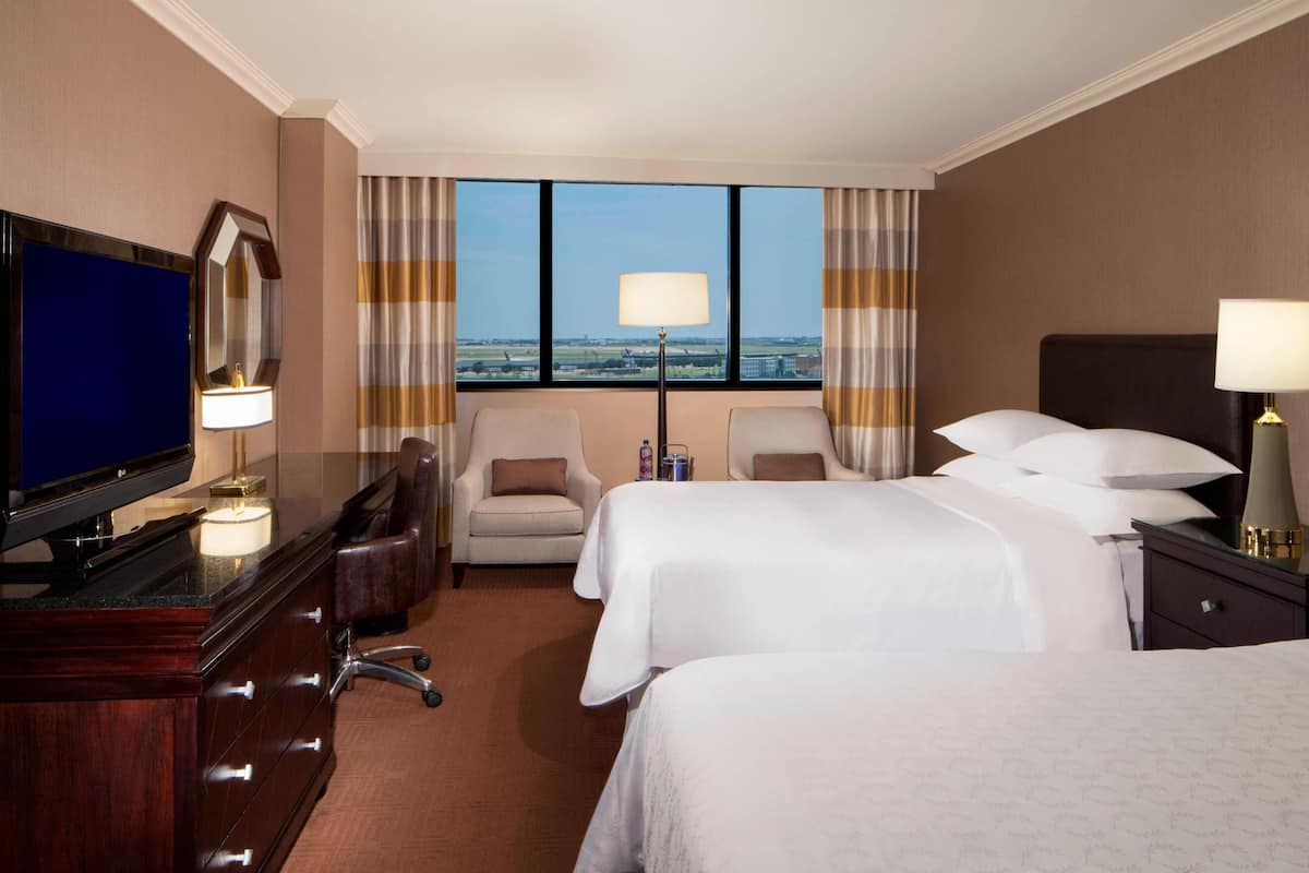 A hotel room with a large TV, two beds, a desk with a computer, and a view of the cityscape.