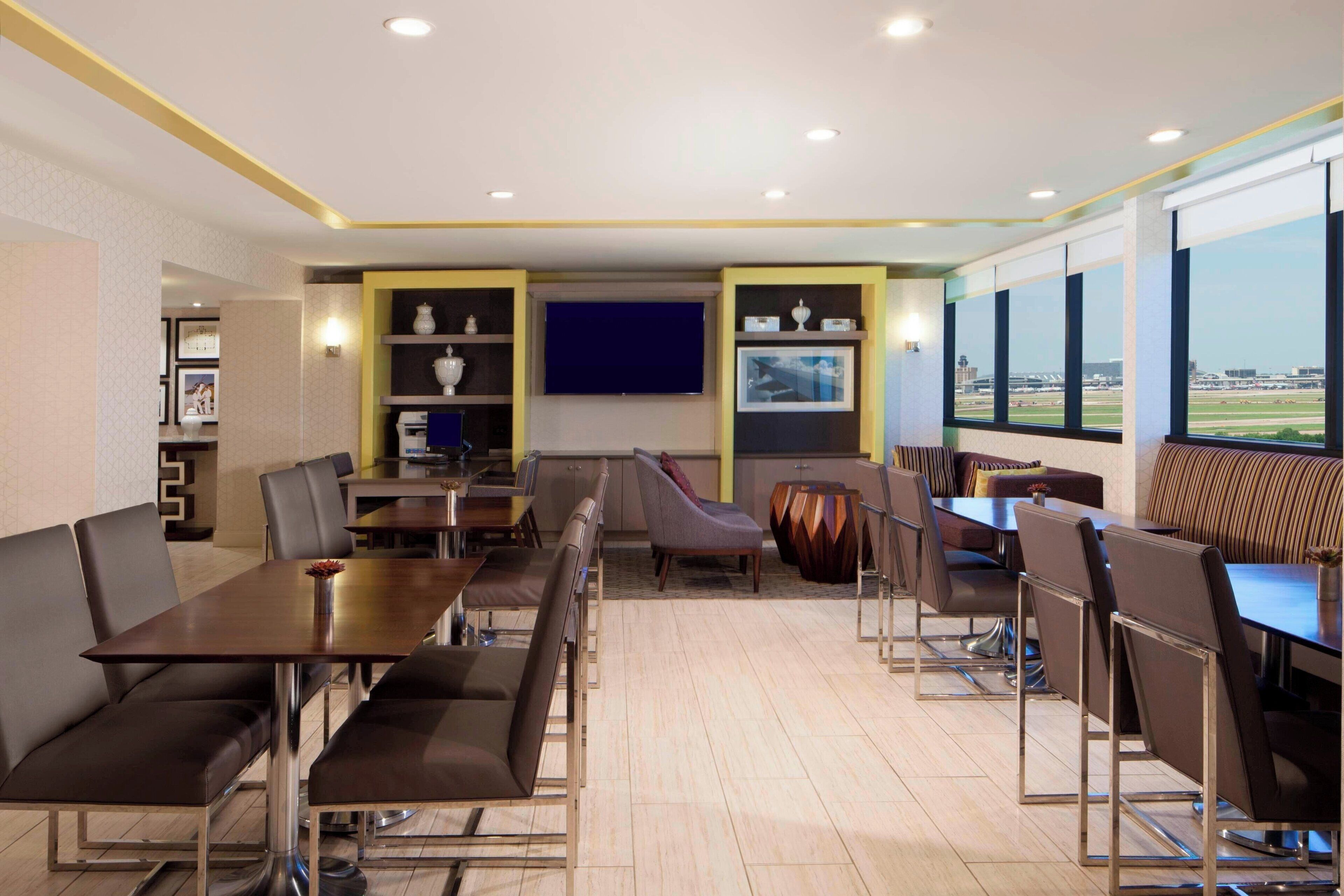 A modern conference room with a long table, multiple chairs, and a sofa. There are shelves with decorative items and a large window with a view of an airport.