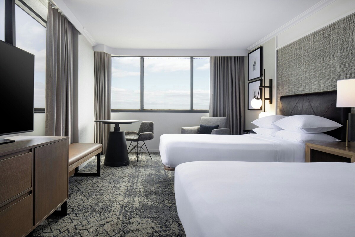 A hotel room with two beds, a desk, a chair, a TV, and a large window with curtains.