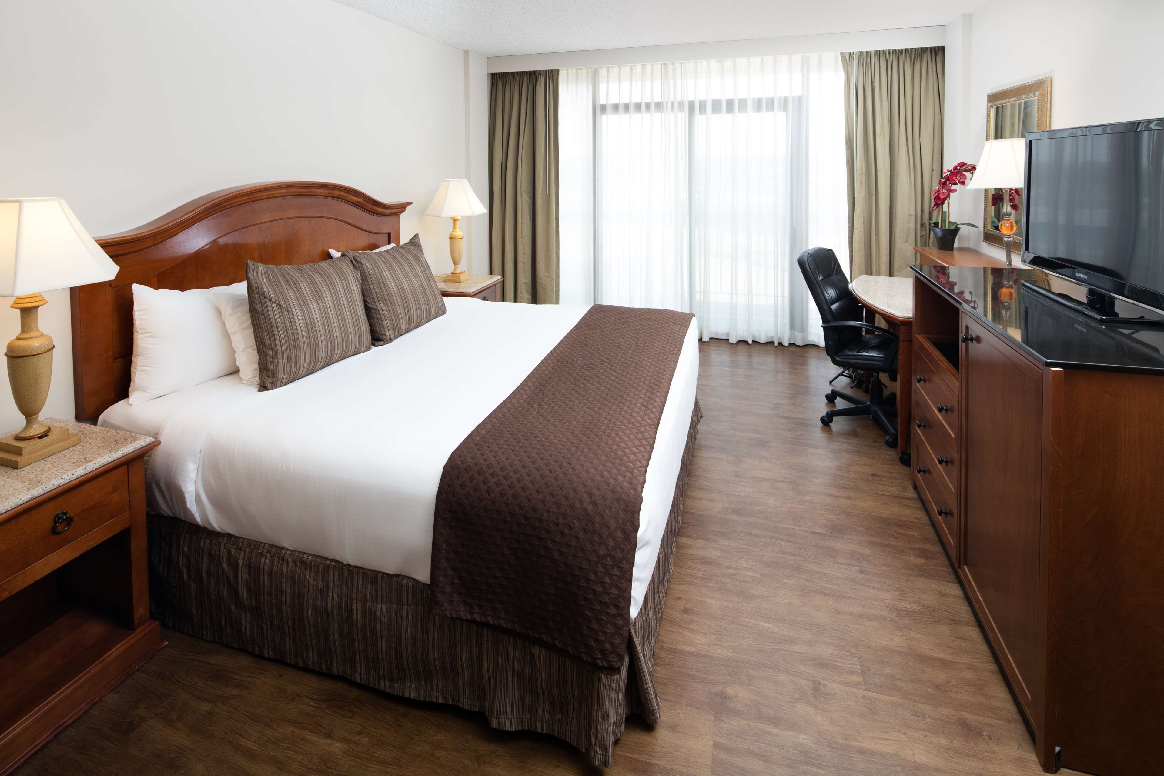 A hotel room with a large bed, bedside tables with lamps, a desk with a chair, a television, and a large window with curtains.