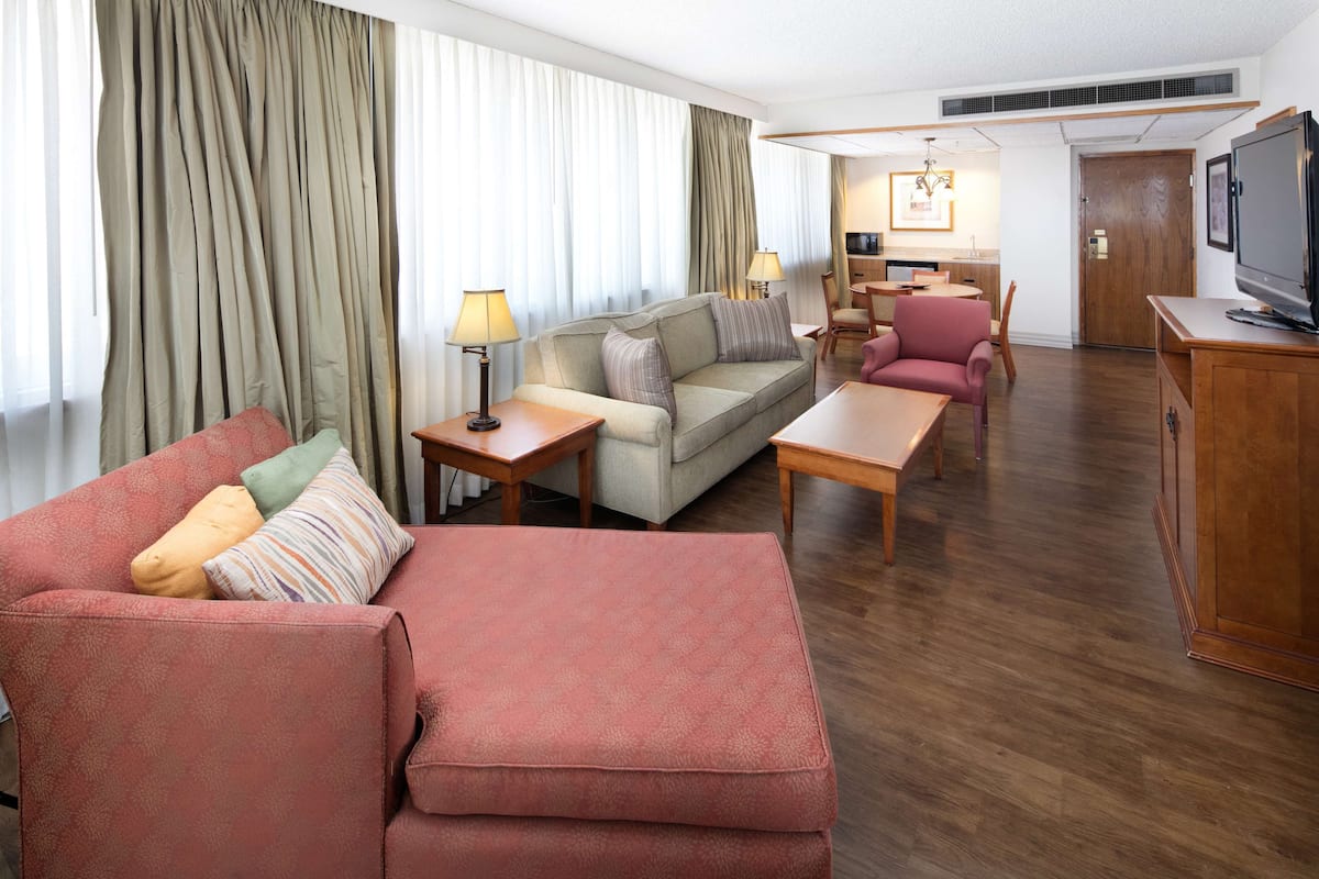 A modern hotel room with a living area featuring a sofa, armchairs, a coffee table, and a TV.