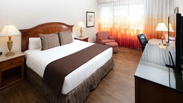 A hotel room with a large bed, two bedside tables with lamps, a desk with a chair, a television, and a window with curtains.