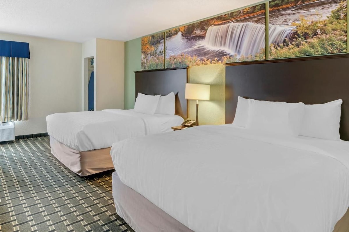 A hotel room with two beds, a large window with curtains, a wall-mounted painting of a waterfall, and a patterned carpet.