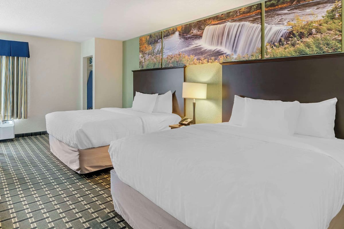 A hotel room with two beds, a large window with curtains, a wall-mounted painting of a waterfall, and a patterned carpet.