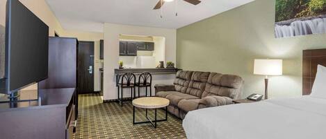A hotel room with a bed, a desk, a television, a ceiling fan, a small table, a sofa, a lamp, and a kitchen area in the background.