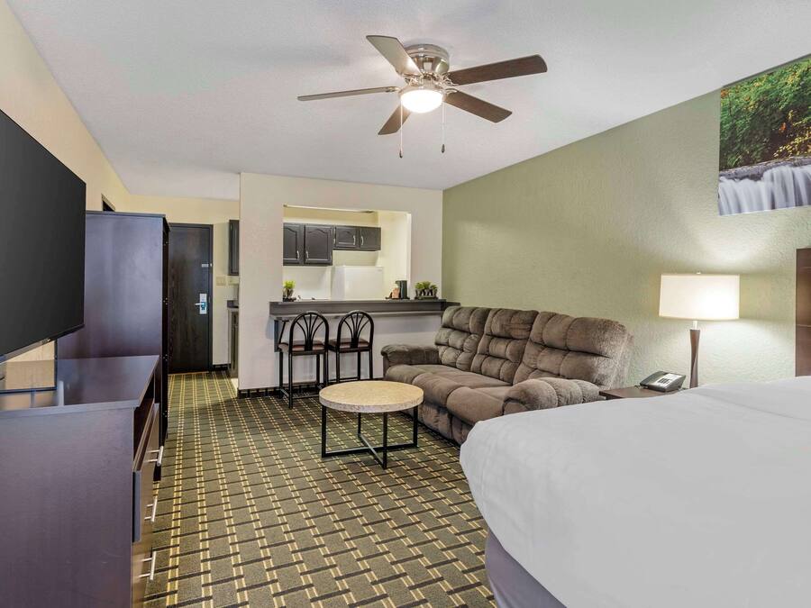 Habitación de hotel con cama, escritorio, televisor, ventilador de techo, una pequeña mesa, sofá, lámpara y una cocina al fondo.