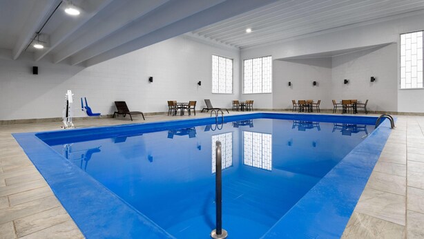 An indoor swimming pool with a blue floor, surrounded by chairs and tables, and large windows on the walls.