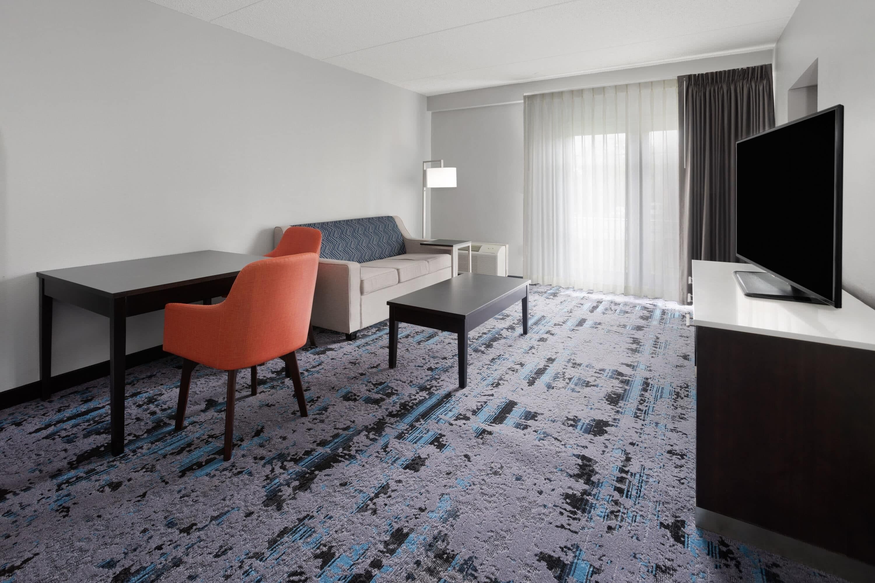 A hotel room with a sofa, two chairs, a desk, a flat-screen TV, a lamp, and a patterned carpet.