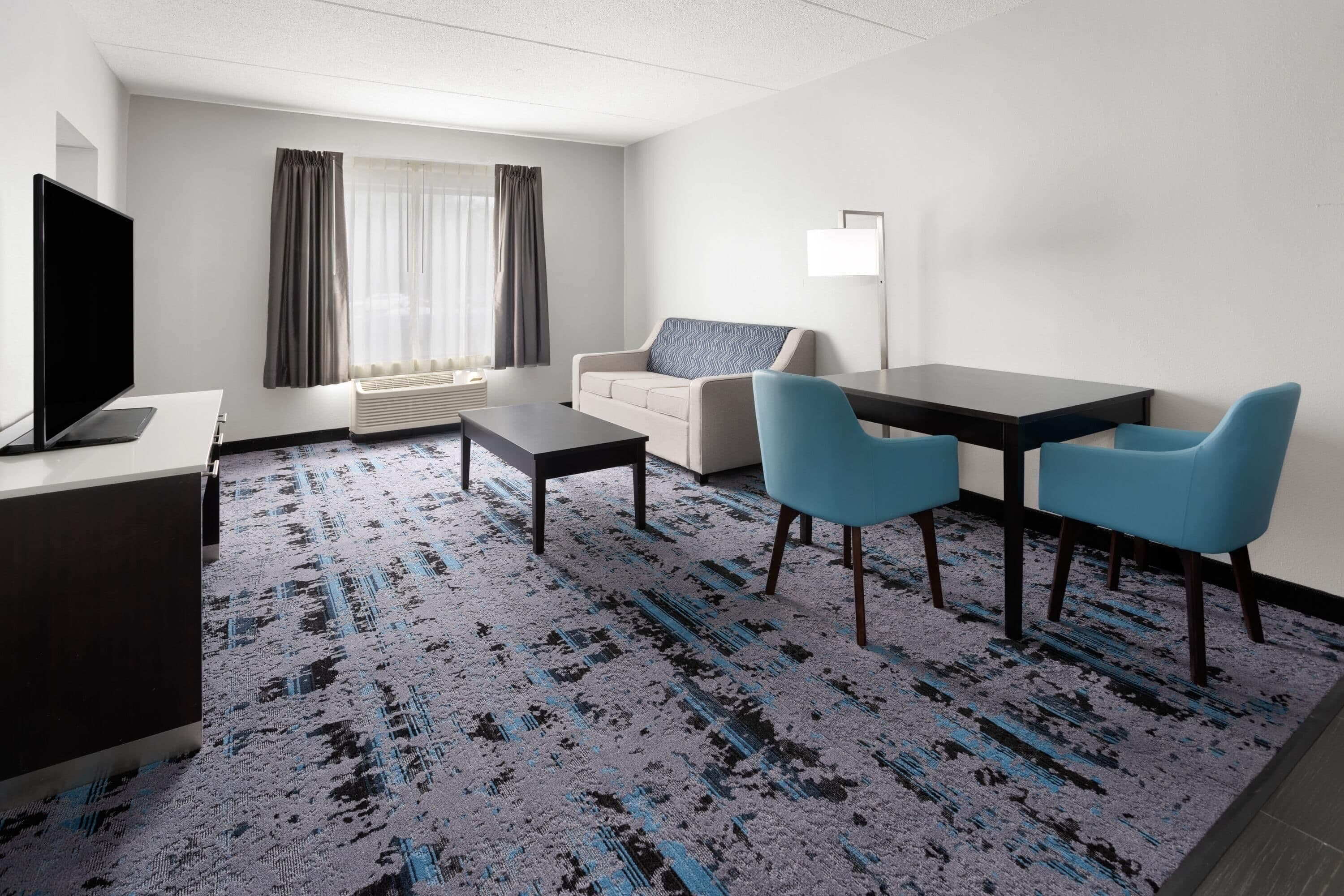 A hotel room with a flat-screen TV, a sofa, a dining table with chairs, and a patterned carpet.