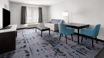 A hotel room with a flat-screen TV, a sofa, a dining table with chairs, and a patterned carpet.