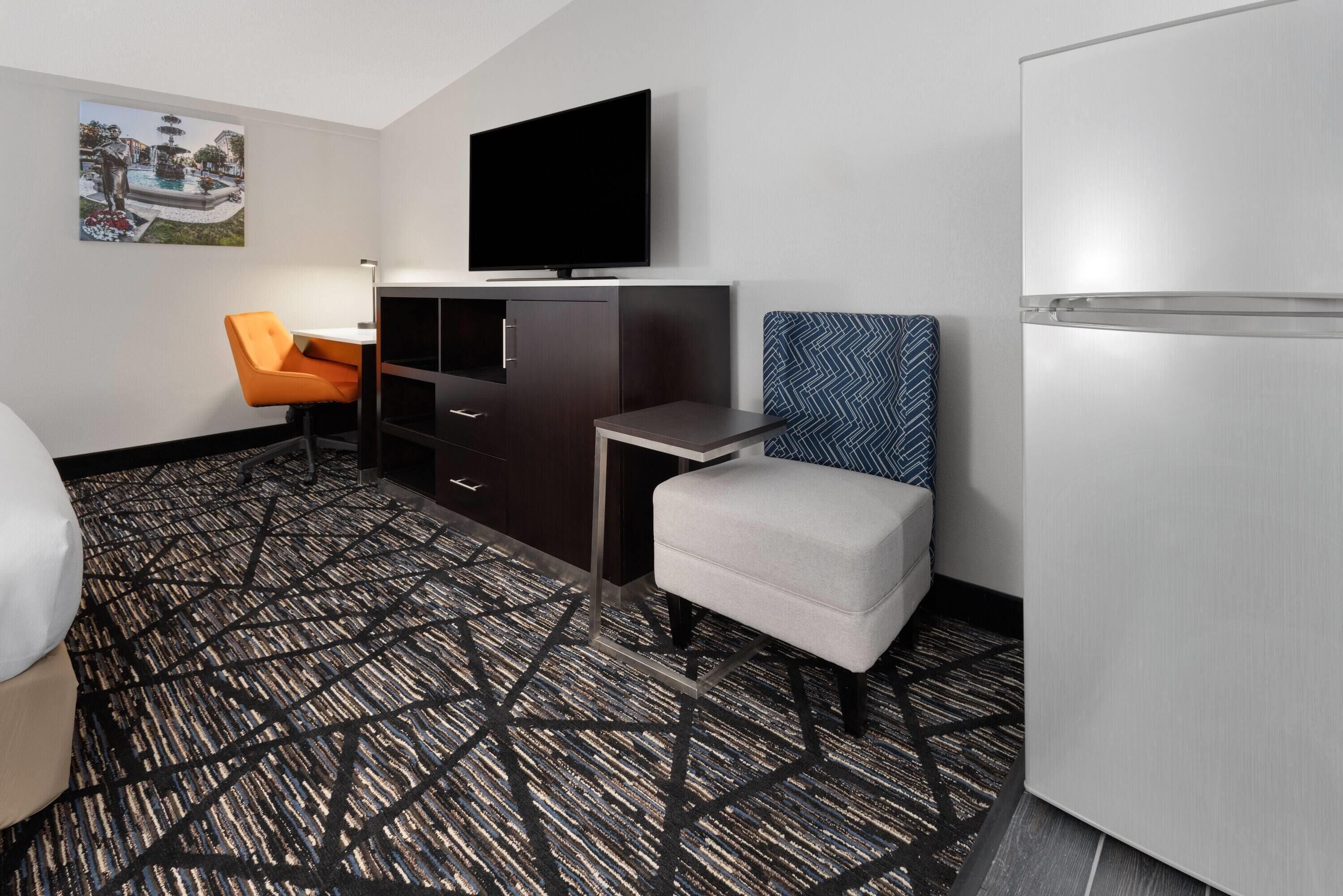 Une chambre d’hôtel équipée d’un lit, d’un bureau, d’une chaise, d’une télévision, d’un réfrigérateur et d’une moquette à motifs.