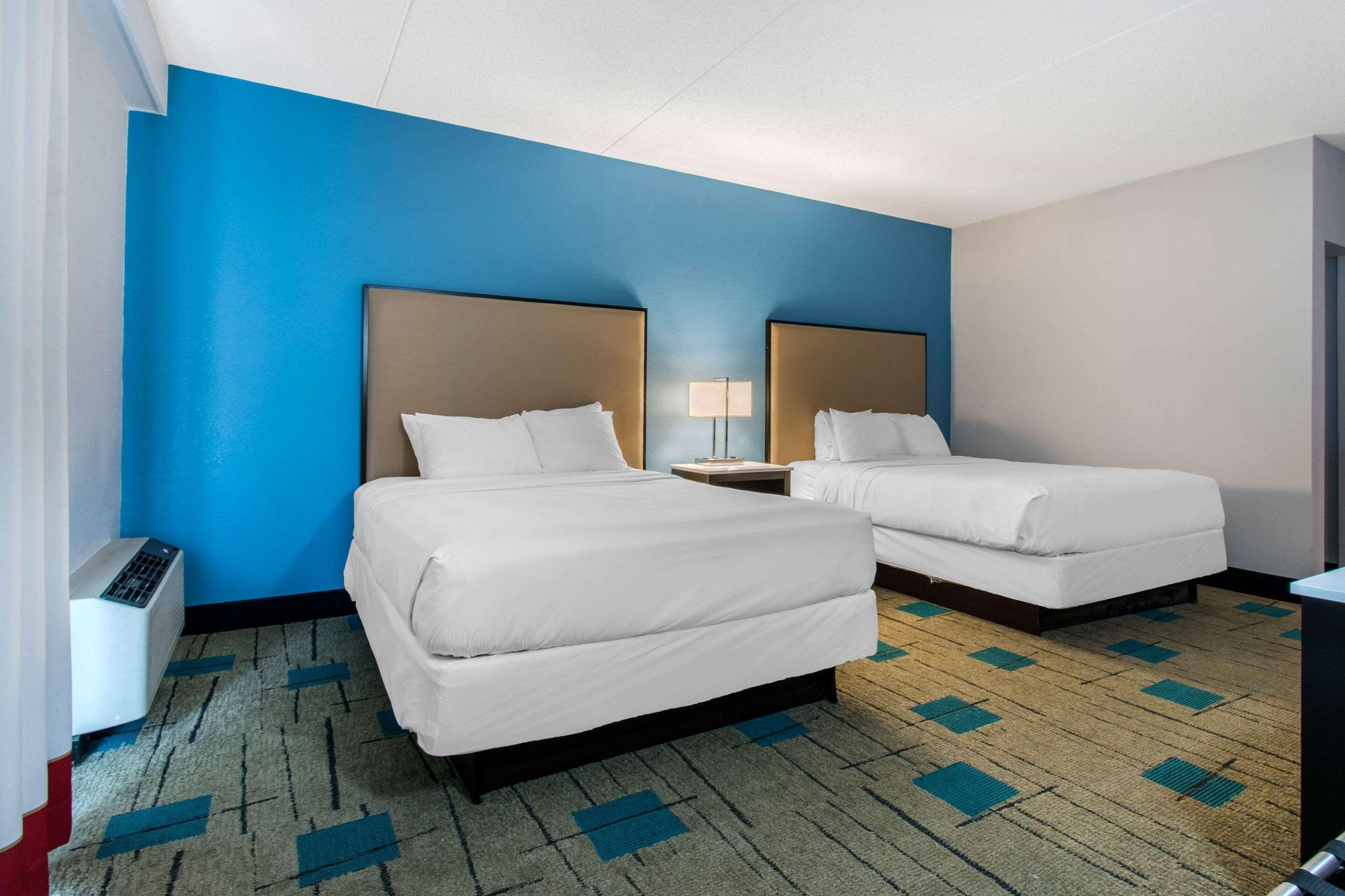 Een hotelkamer met twee bedden, een nachtkastje met lamp, een heater en een vloerkleed met een geometrisch patroon.