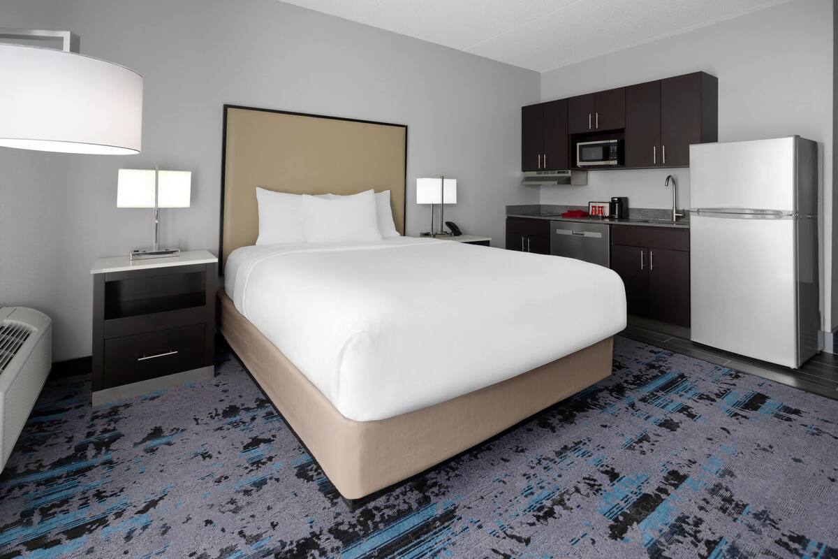 Ein Hotelzimmer mit einem großen Bett, einem Nachttisch mit Lampe, einem kleinen Schreibtisch, einer Mikrowelle, einem Kühlschrank und einer Küchenzeile.