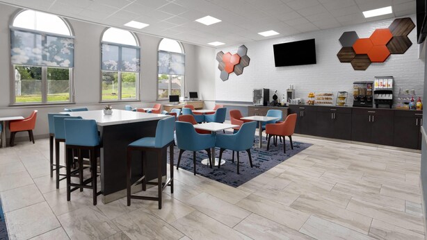 A modern dining area with high tables and blue chairs, a kitchen area with a coffee machine, and a large window with a view of the outdoors. - Hawthorn Extended Stay by Wyndham Chambersburg (Chambersburg)