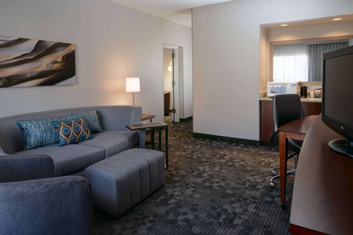 A hotel room with a grey sofa, a small table, a TV, and a kitchenette area.