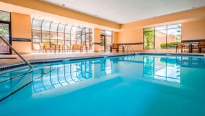 A swimming pool with clear blue water, surrounded by a sunlit indoor area with large windows, a few chairs, and a table. - Sonesta Select Kansas City South Overland Park (Kansas City)