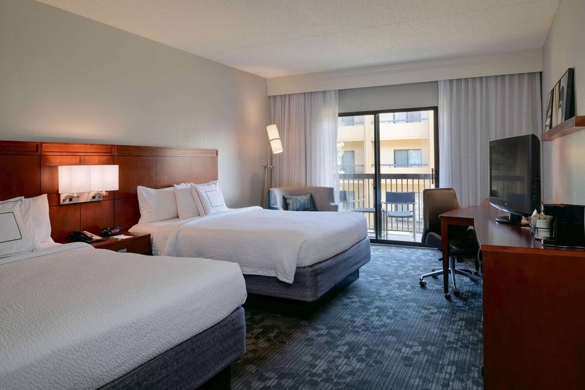 A hotel room with two beds, a desk, a TV, and a balcony with a view of the building.