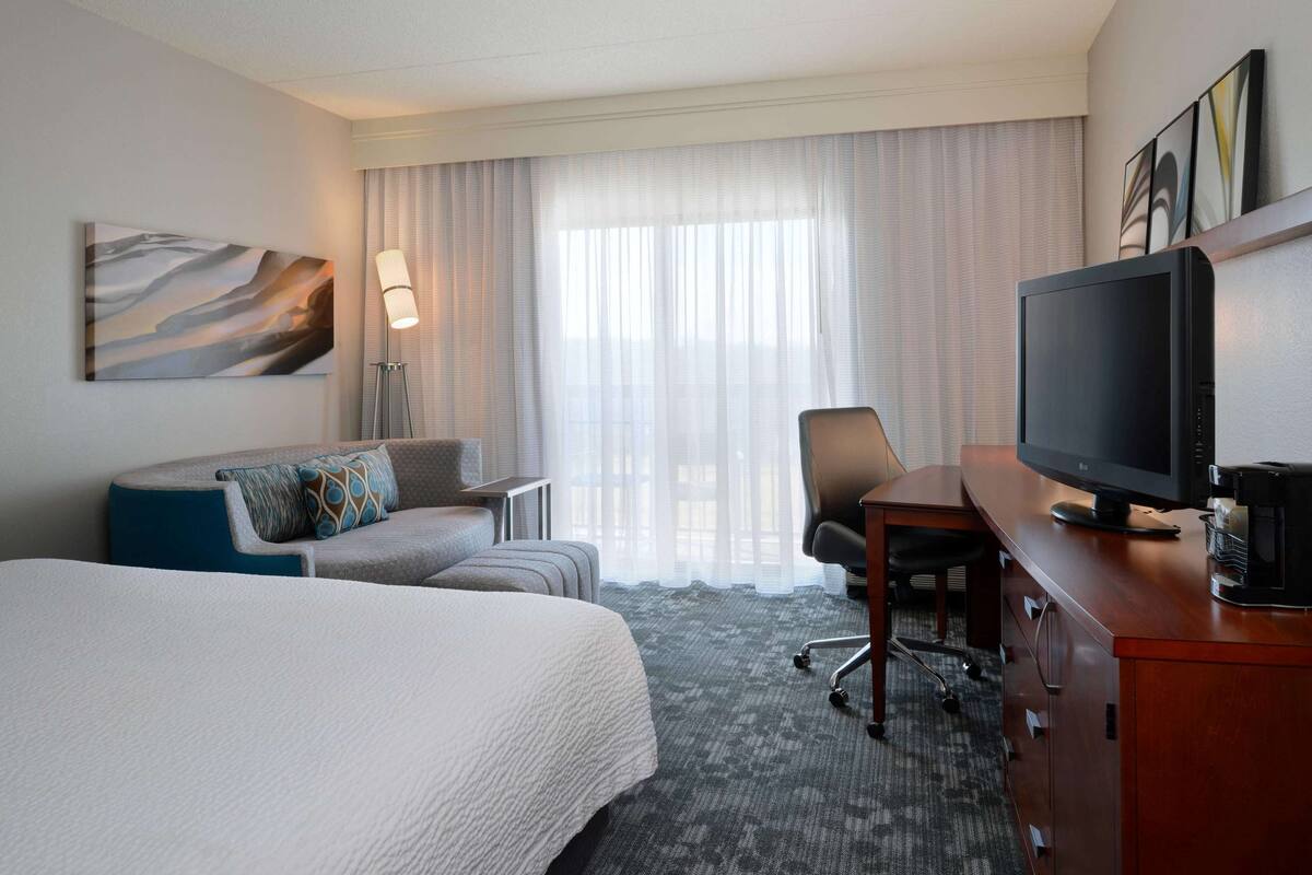 A hotel room with a large bed, a desk with a television, a chair, and a lamp.