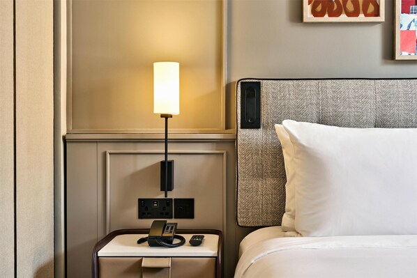 A hotel room with a bed, a bedside table with a lamp, a phone, and a wall-mounted control panel.