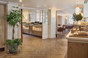 Um saguão moderno de hotel com piso de madeira, balcão de recepção, área de refeições com mesas e cadeiras, e cozinha com panelas e utensílios.