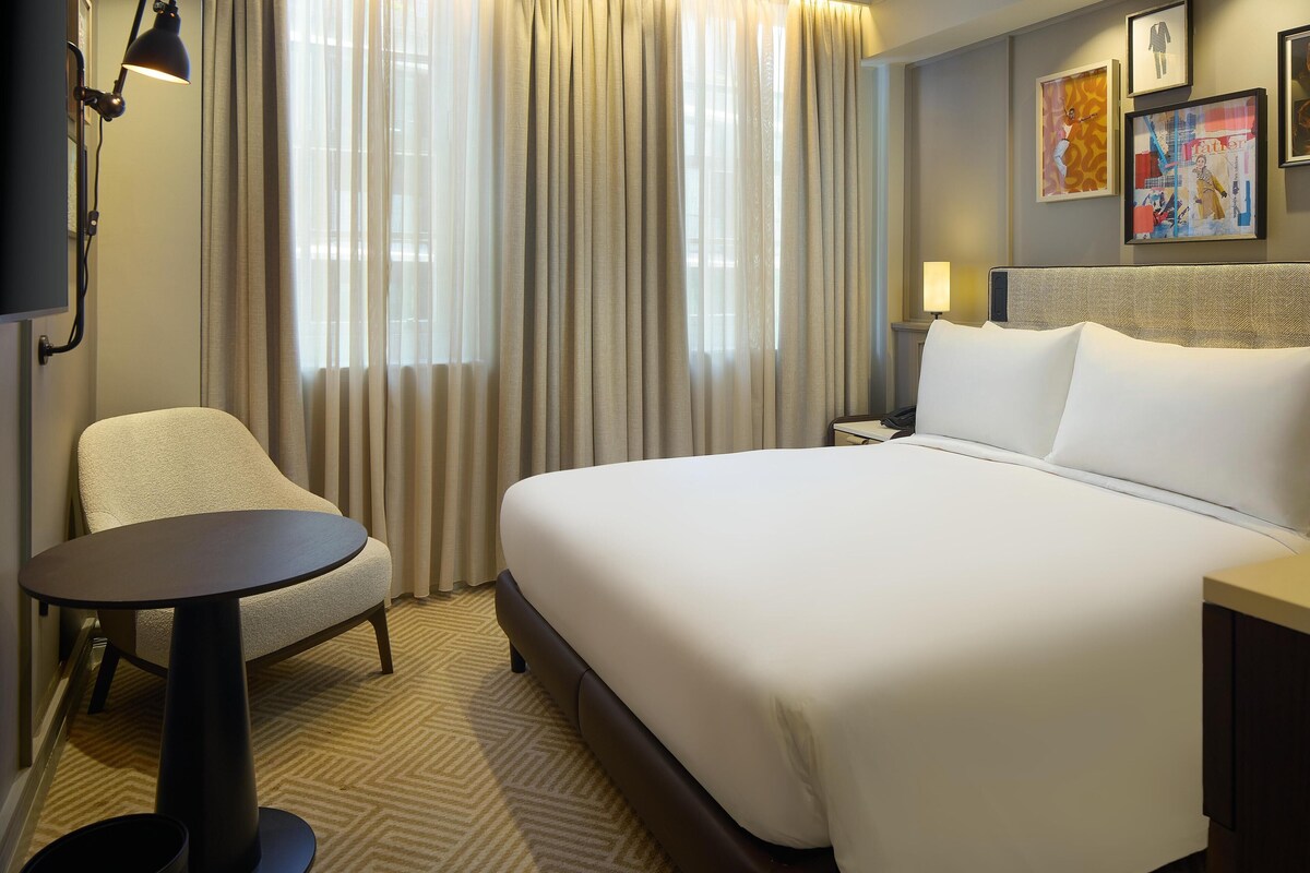 Una habitación de hotel con una cama, una silla, una mesita, una mesita de noche y una lámpara.