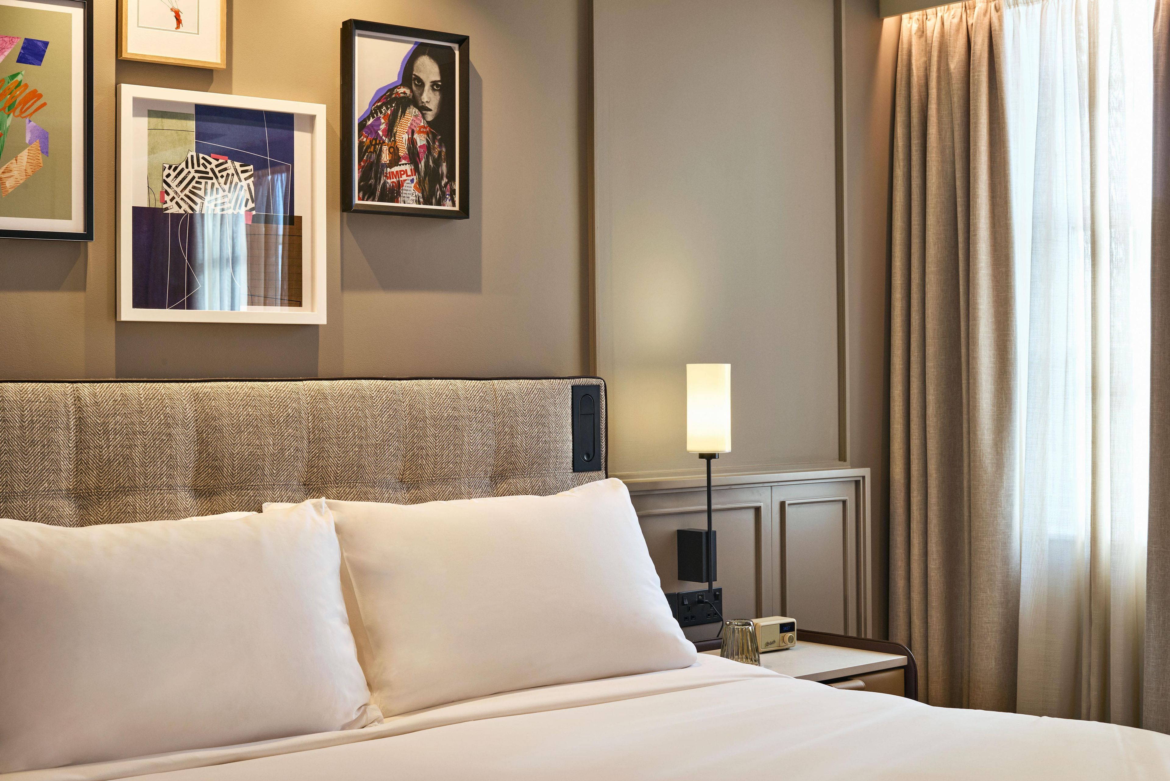 Una habitación de hotel con una cama bien hecha, una mesita de noche con lámpara y cuadros en la pared.