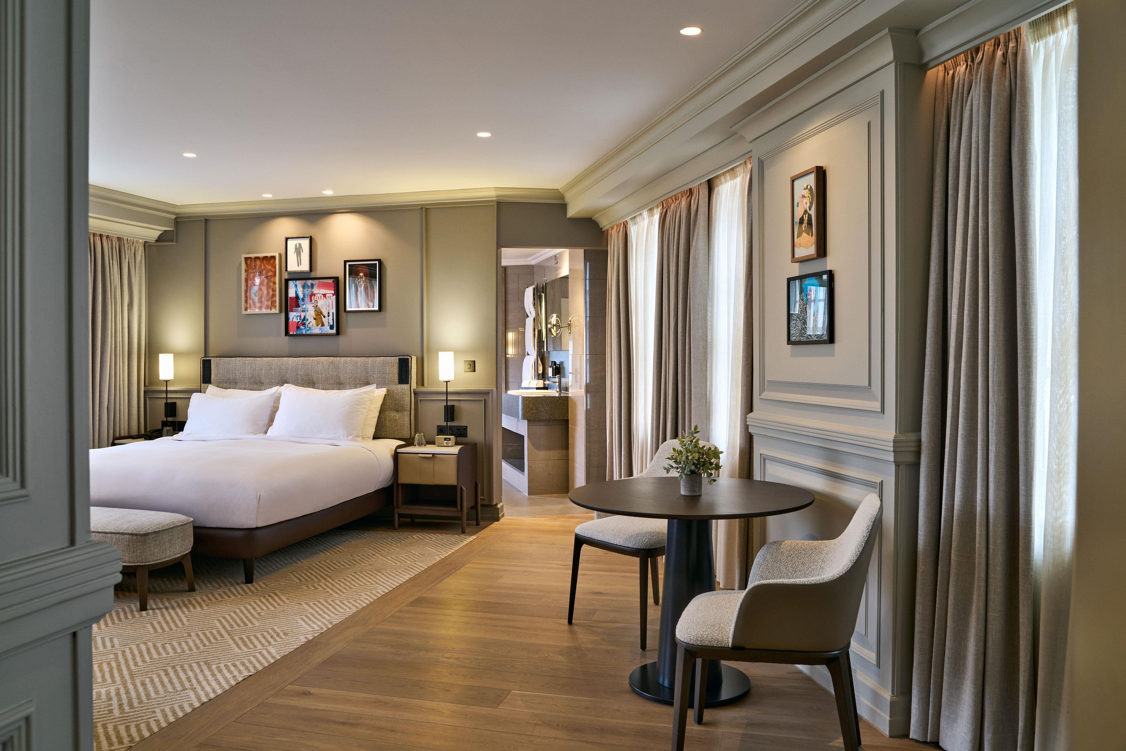 A hotel room with a large bed, a small table with chairs, a desk, and a bathroom visible through an open door.