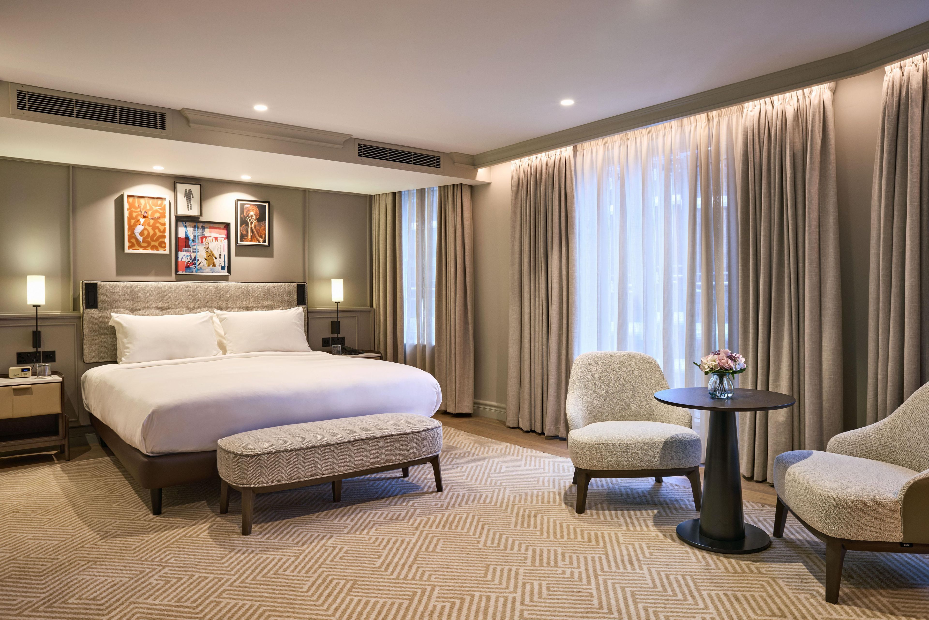 Una habitación de hotel con una cama grande, dos mesitas de noche, una pequeña mesa redonda con un florero y dos sillones, además de una alfombra estampada.