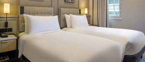 Habitación de hotel con dos camas, una mesita de noche con lámpara, un cuadro en la pared y una ventana con cortinas.
