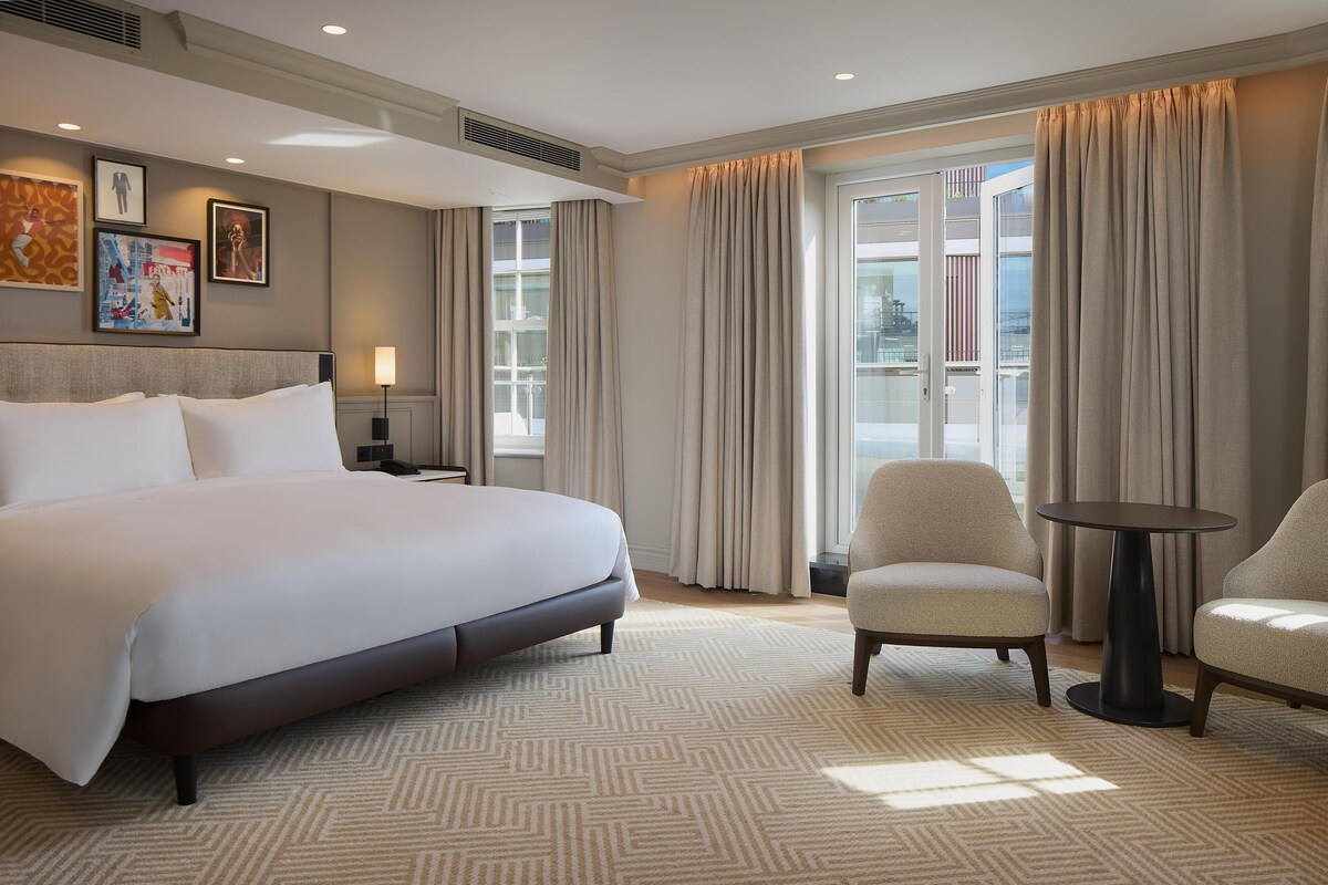 Habitación de hotel con una cama grande, dos sillones, una mesita y un balcón con cortinas.