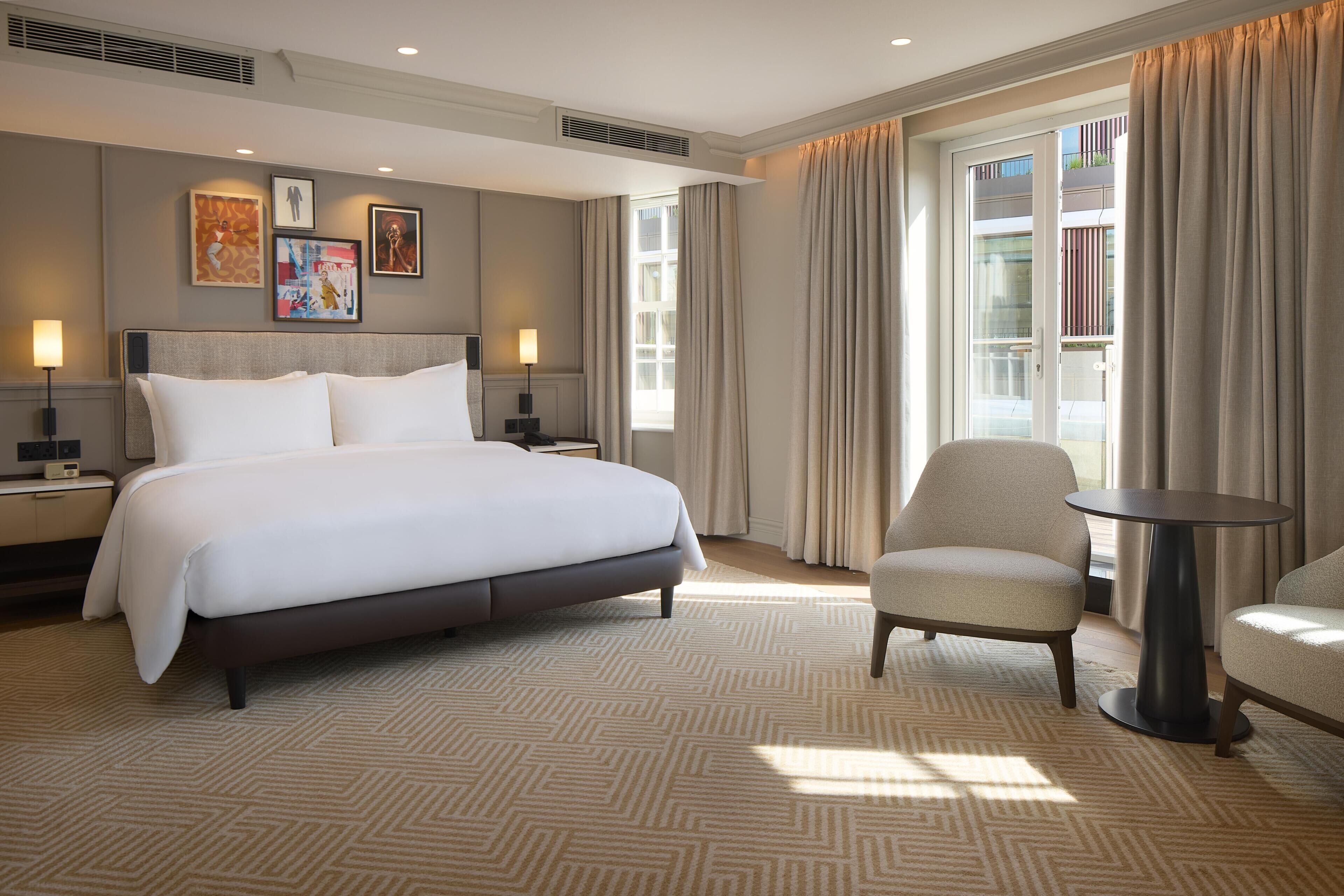 A hotel room with a large bed, two bedside tables, a chair, a small round table, and a large window with curtains.