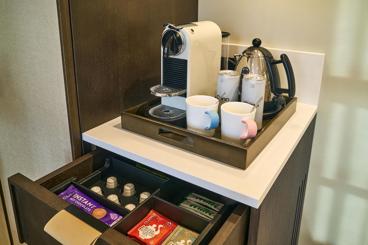 Una estación de café con una cafetera, tazas y una bandeja. Hay un cajón debajo con cápsulas de café y una caja de café instantáneo.