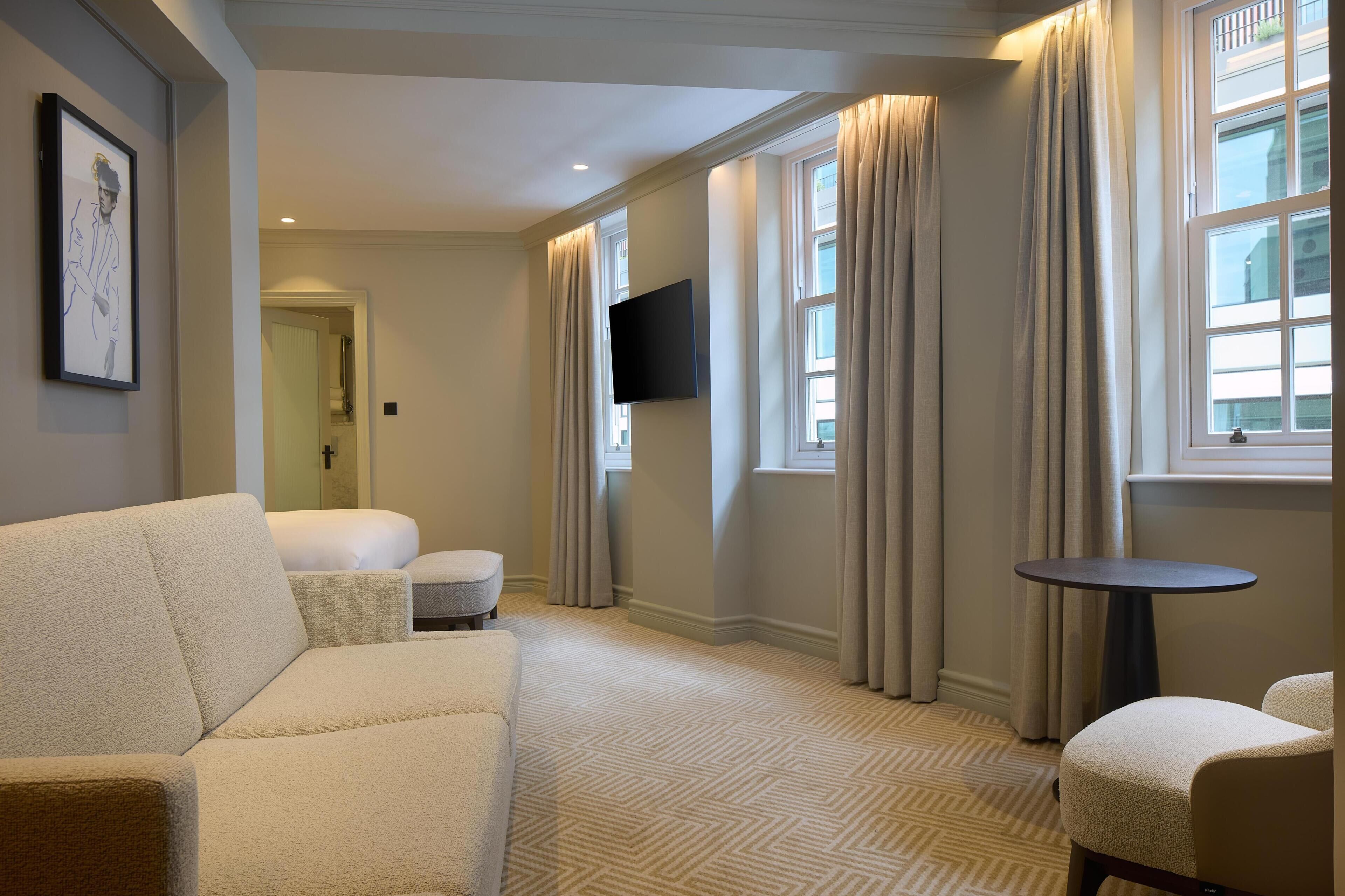 A hotel room with a sofa, a small round table, a flat-screen TV mounted on the wall, and a window with curtains.