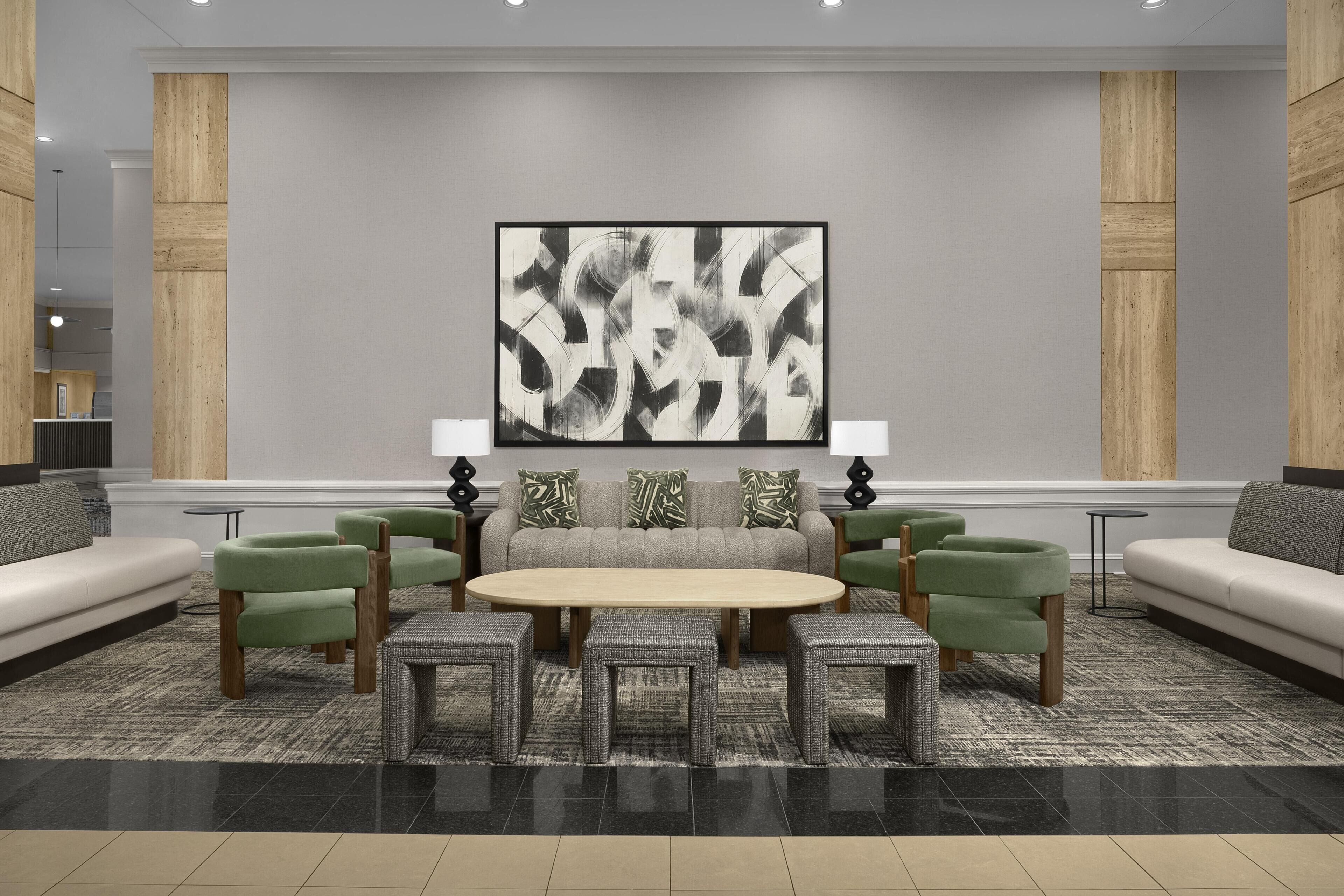 A modern lounge area with a central round table, green upholstered chairs, a grey sofa, and a large abstract painting on the wall.