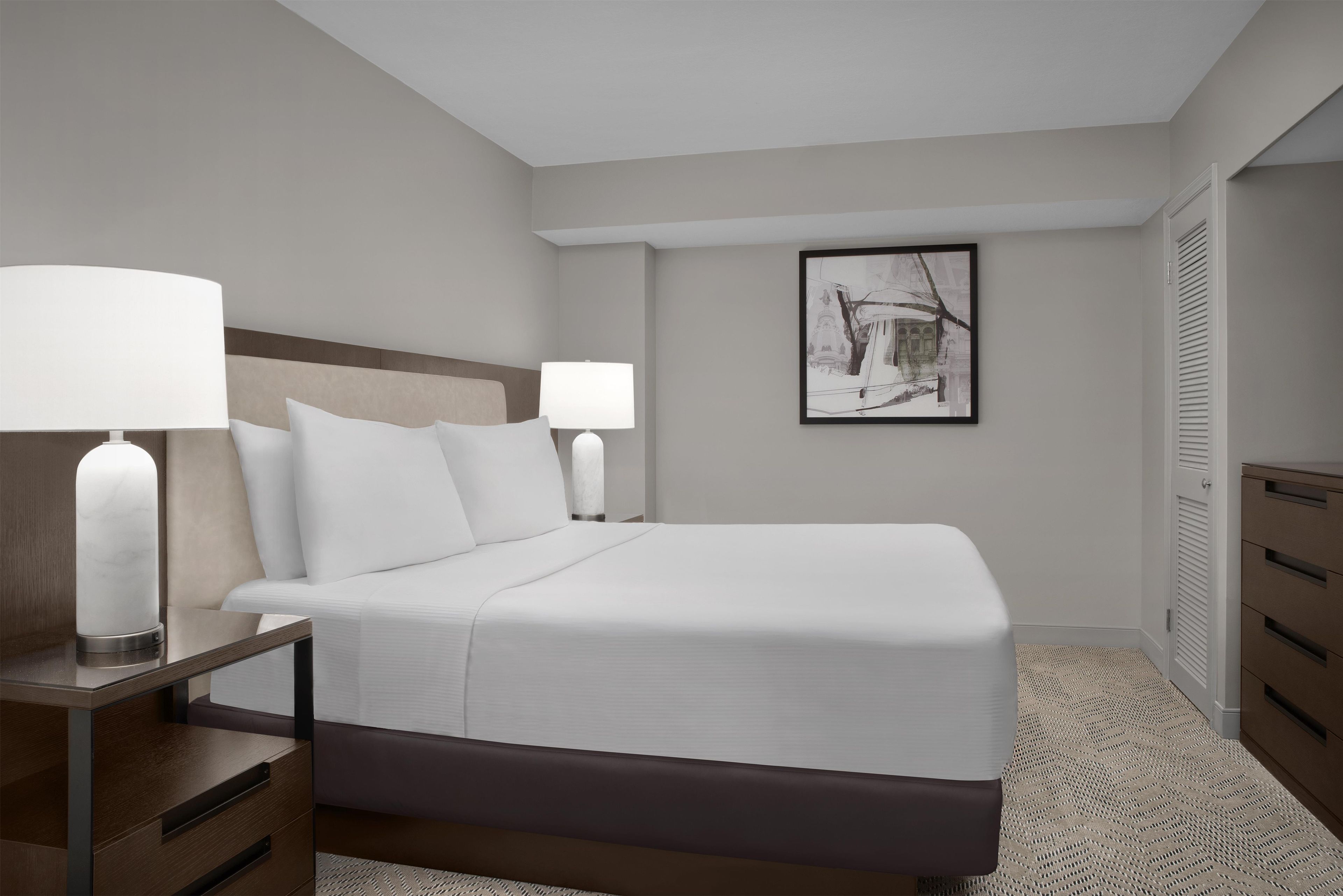 A hotel room with a large bed, two bedside tables with lamps, a framed artwork on the wall, and a wardrobe.