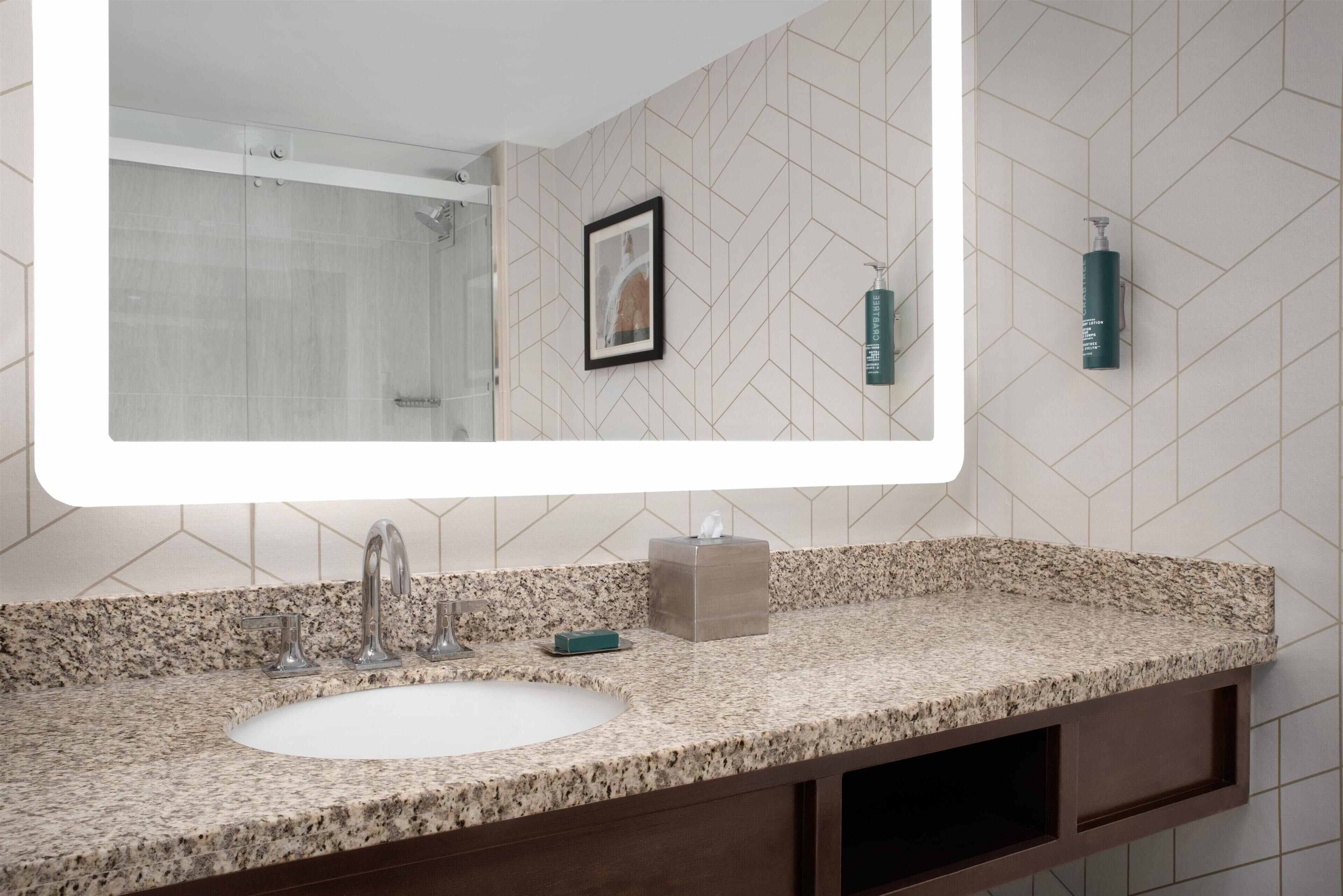 A bathroom with a large mirror, granite countertop, sink, and a framed picture on the wall.
