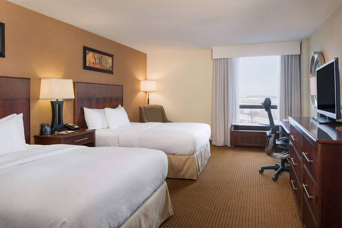 A hotel room with two beds, a desk with a TV, a chair, and a window with a view.