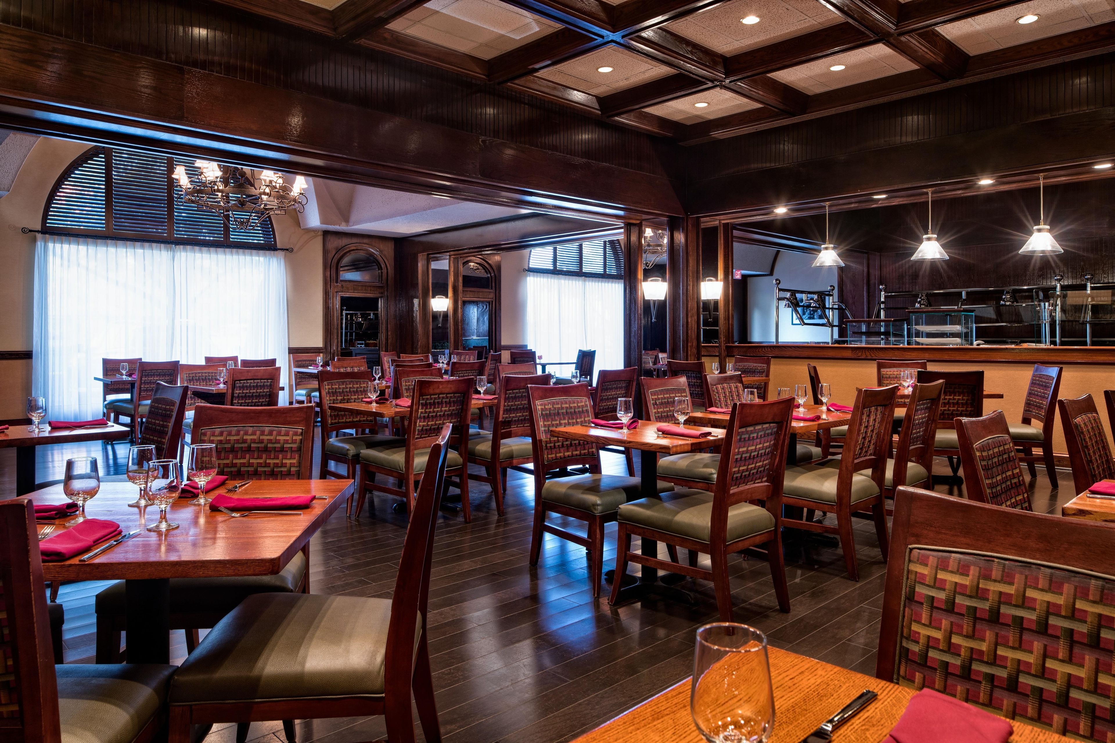 Un restaurante con mesas y sillas de madera, ventanas amplias y luces colgantes.