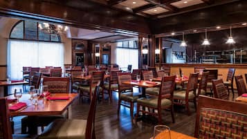 Un restaurante con mesas y sillas de madera, ventanas amplias y luces colgantes.