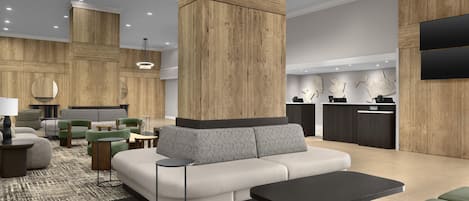 A modern hotel lobby with a large sofa, green ottomans, a black coffee table, and a reception desk with a flat-screen TV.
