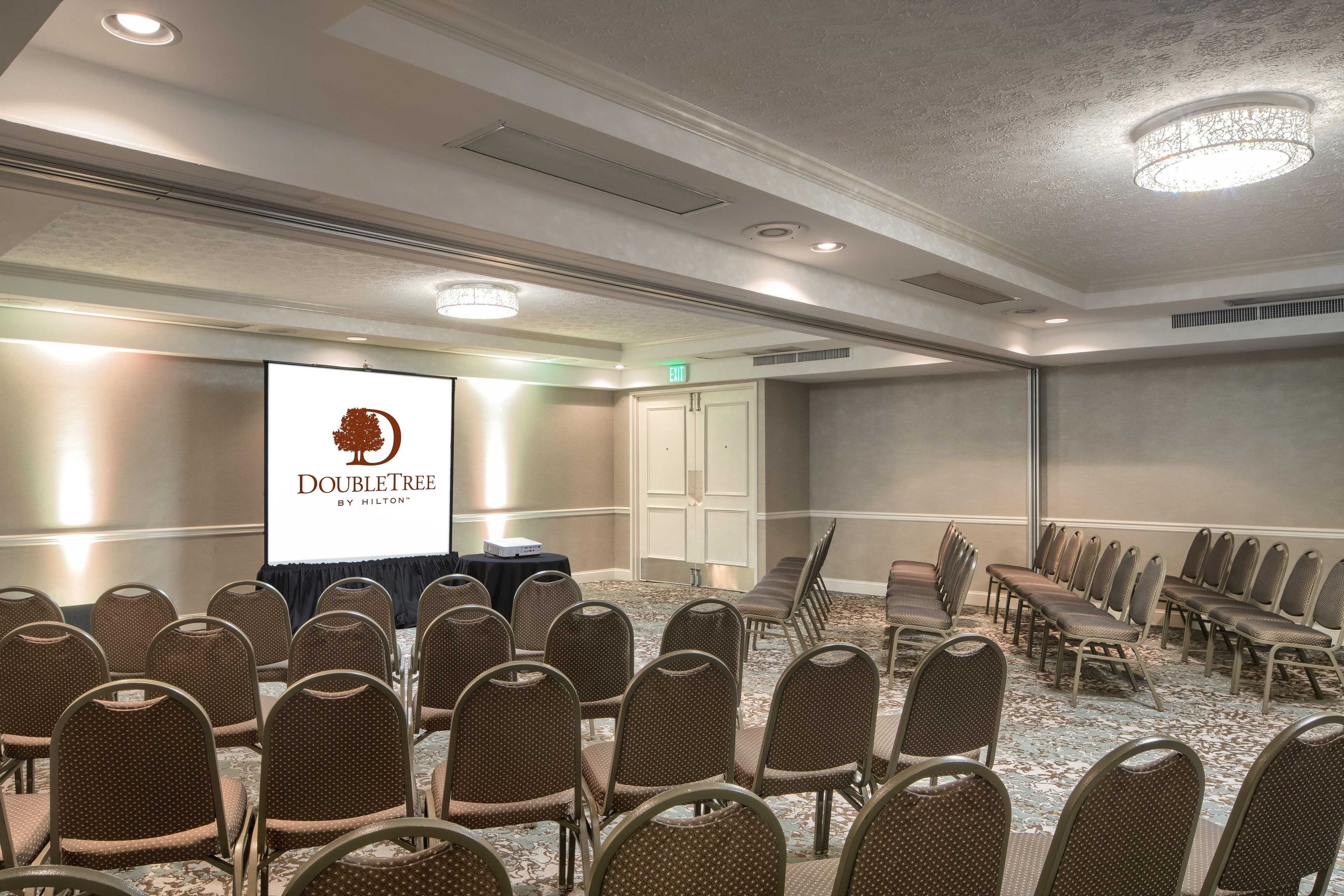 Un salón de conferencias con filas de sillas, una pantalla de proyector mostrando un logotipo y una puerta de salida.