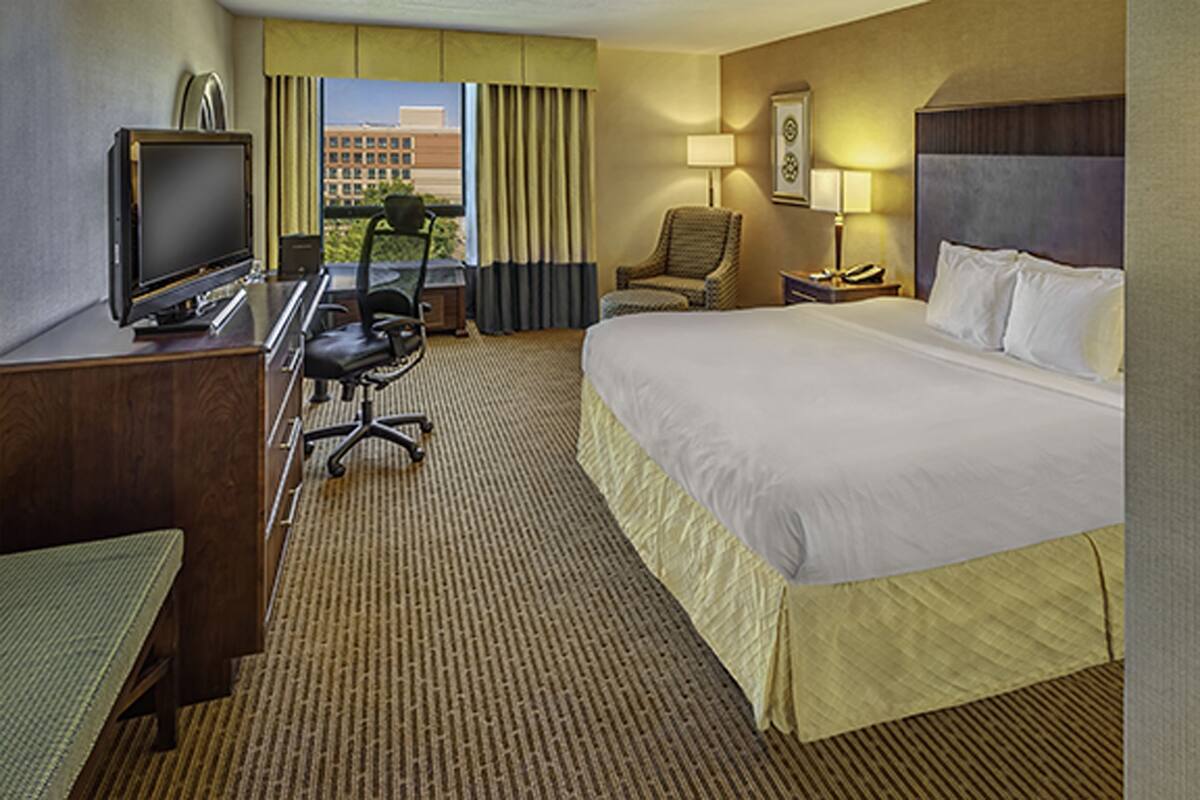 A hotel room with a bed, desk, chair, television, lamp, and a view of the cityscape.