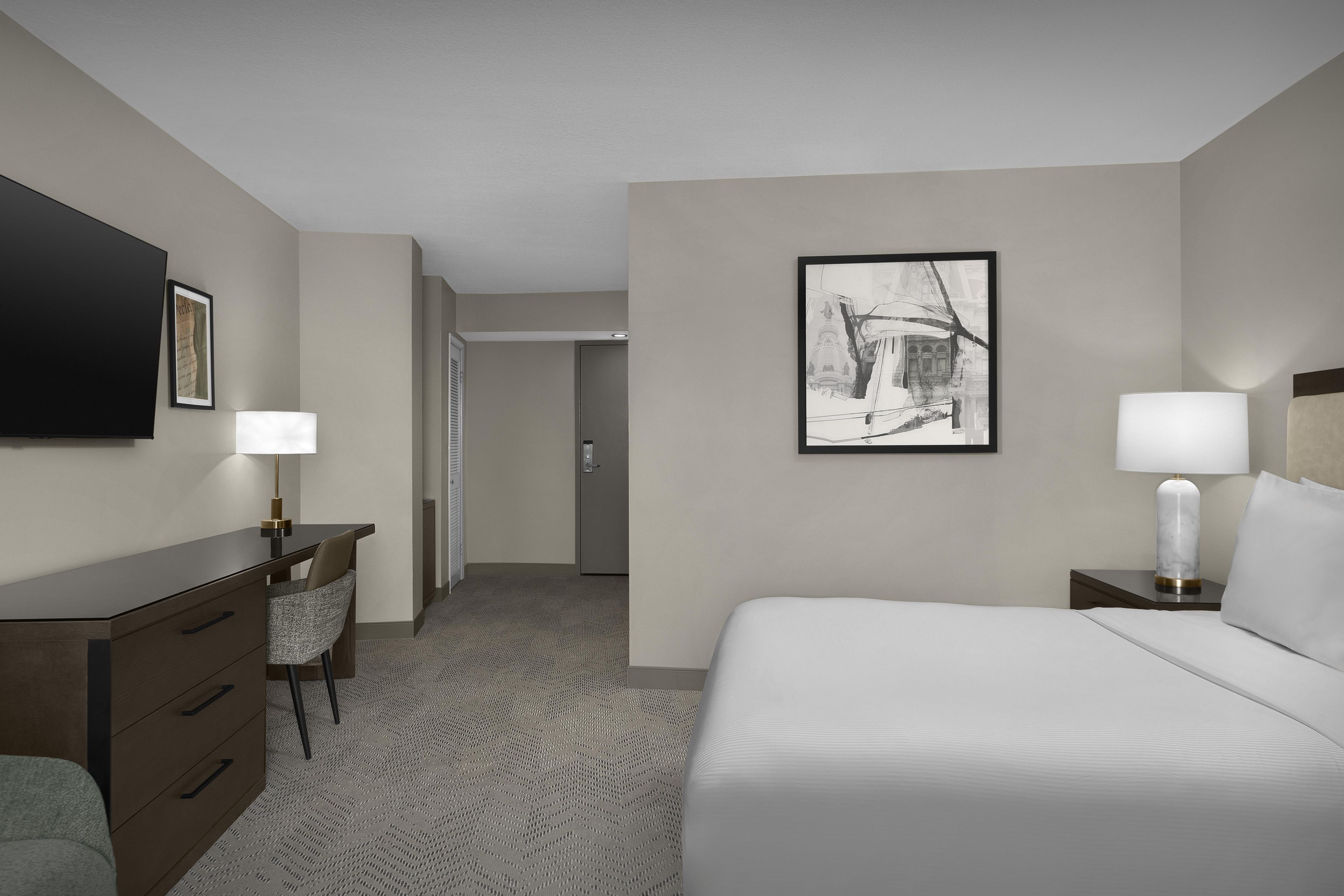 Une chambre d’hôtel avec un lit, un bureau, une chaise, une télévision et un tableau au mur.