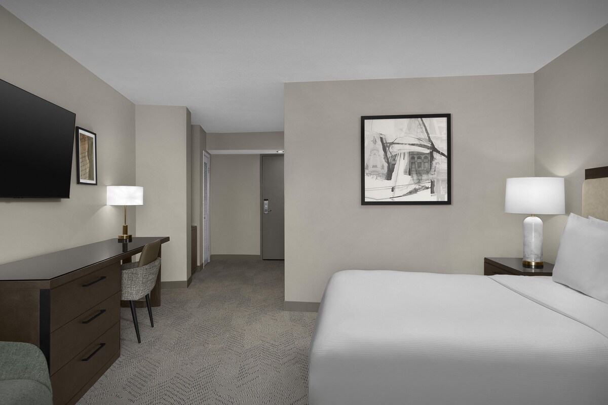 Una camera d'albergo con un letto, una scrivania, una sedia, una televisione e un quadro appeso al muro.