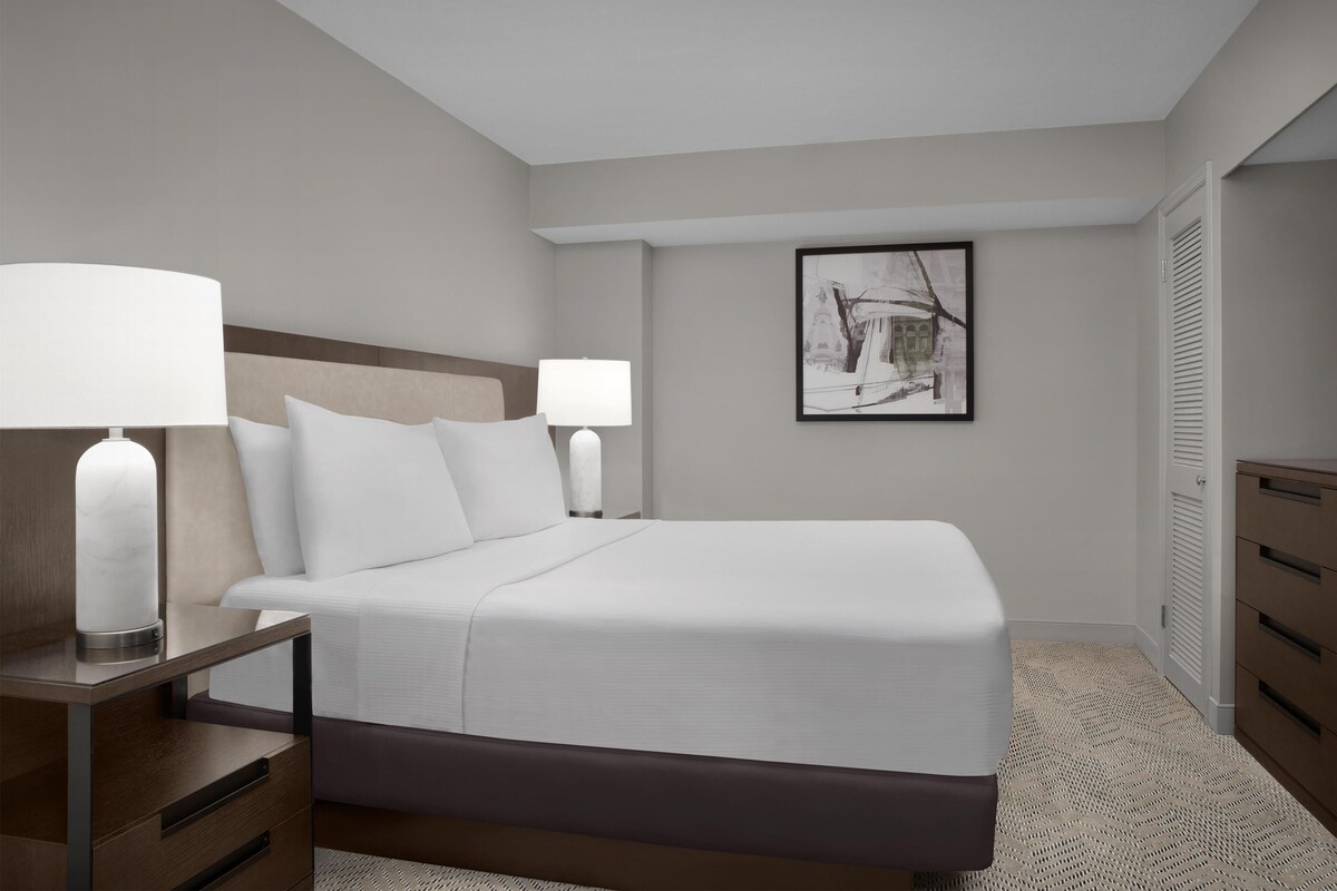A hotel room with a bed, two bedside tables with lamps, a framed picture on the wall, and a wardrobe.