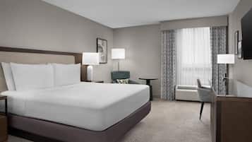 Quarto de hotel com uma cama grande, duas mesinhas de cabeceira, uma cadeira, uma escrivaninha, um abajur, um quadro na parede e uma janela com cortinas.